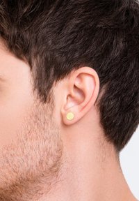 Goldene Metallstecker mit strukturierter Oberfläche, runde Form, am Ohrläppchen getragen, vor einem hellen Hintergrund. Kurzes Haar und Bart sichtbar.