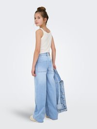 Vaqueros de pierna ancha en azul claro con cintura alta, combinados con una camiseta sin mangas blanca; chaqueta vaquera colgada sobre un brazo. Textura suave, ajuste informal.