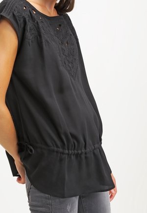 Kobieta nosząca luźną czarną bezrękawnikową bluzkę z haftowanymi detalami i marszczoną talią, w połączeniu z szarymi postrzępionymi jeansami.