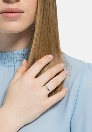 Giovane con pelle chiara e capelli biondi lisci indossa un anello di diamante solitario argentato sull'anulare della mano sinistra.