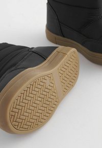 Bottes isolées noires avec un dessus en synthétique lisse, présentant une semelle en caoutchouc texturé de couleur tan avec une adhérence à motif. Coutures accentuées visibles.