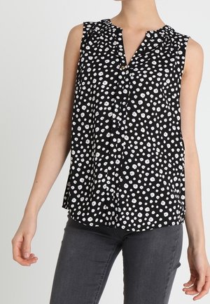 Mouwloze zwarte blouse met witte abstracte stippen, geplooide halslijn, gedragen met donkergrijze spijkerbroek.