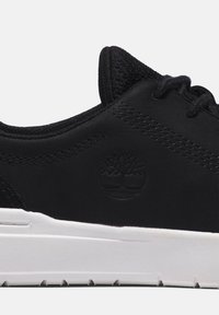 Black sneaker with a textured upper, featuring a logo in relief, white sole with grip patterns, and contrasting stitching details.