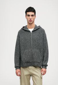 Sivý zipsový mikina s textúrovanou pleteninou, ktorá má kapucňu, predné vrecká a ribbovanými manžetami, v kombinácii s svetlo béžovými cargo nohavicami.