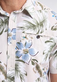 Camisa de manga corta de algodón ligero con un fondo crema decorado con hojas de palma verdes y patrones florales azules; incluye un bolsillo en el pecho con botón.