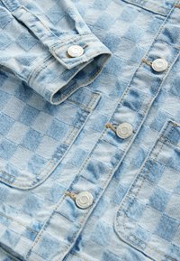 Veste en denim bleu clair avec un motif à carreaux, deux poches sur le devant, des boutons en argent et des détails de couture visibles.