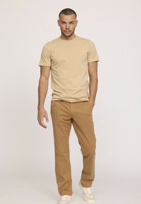 T-shirt en coton beige avec un col rond, accompagné de chinos beiges. Modèle portant des baskets claires. Tenue simple et décontractée.
