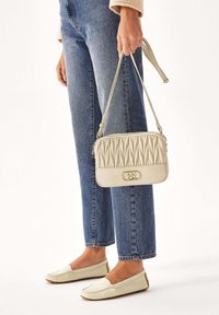 Borsa a tracolla beige chiaro trapuntata con forma rettangolare e chiusura a zip, tenuta da una persona che indossa jeans blu e scarpe basse chiare.