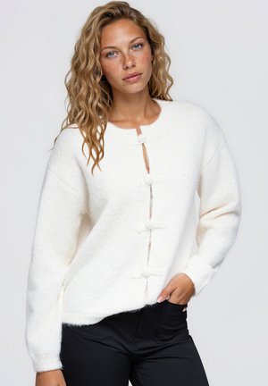 Une femme aux taches de rousseur porte un cardigan blanc duveteux à brandebourgs et un pantalon noir, sur un fond clair uni.