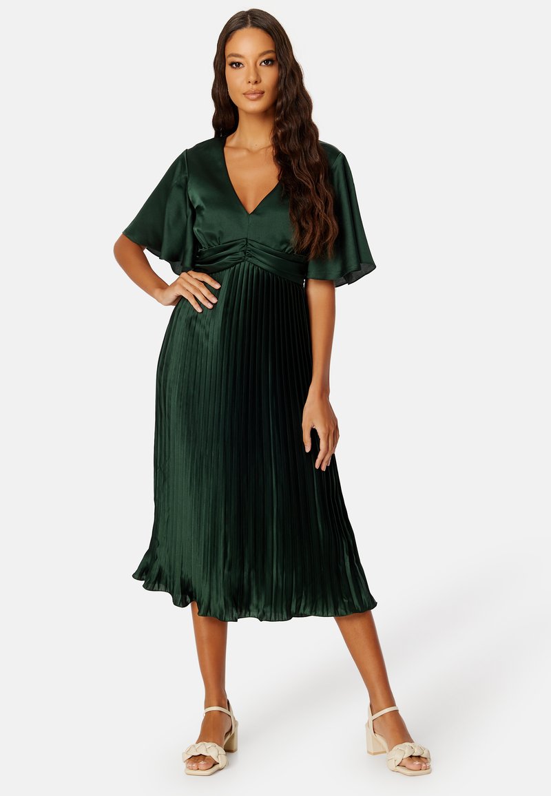 Robe plissée en satin vert avec un décolleté en V profond, des manches courtes volantées et un ourlet fluide. Complétée par des sandales à talons bloc à lanières beiges.