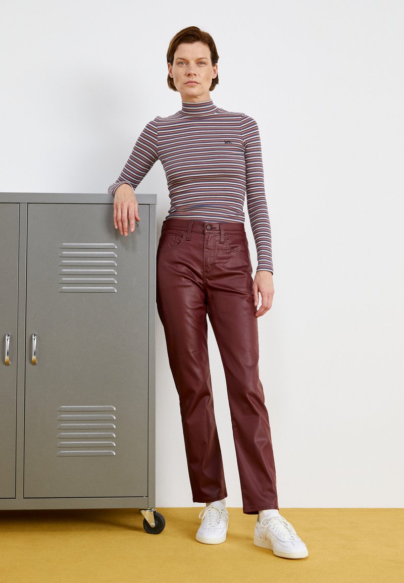 Burgunderfarbene Lederhosen, figurschmeichelndes Design. Weiche, glatte Textur. Langärmeliges, gestreiftes Oberteil mit Stehkragen. Weiße Sneaker. Graues Aufbewahrungselement im Hintergrund.
