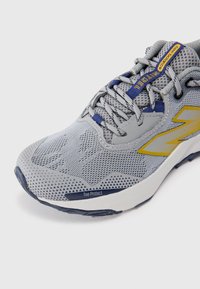 Chaussure de running pour homme avec une tige en mesh gris, des accents bleus, un capuchon de protection pour les orteils et une semelle en caoutchouc texturée pour une meilleure adhérence et durabilité.
