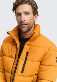 Oranje gewatteerde jas met hoge kraag, zwarte rits en gestikte ontwerpen; gedragen over een zwart shirt met een borstzak.