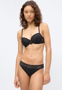 Γυναίκα με σγουρά μαλλιά που φοράει μαύρο σουτιέν από δαντέλα και αντίστοιχο εσώρουχο με δαντελένιο τελείωμα, στέκεται μπροστά σε απλό ανοιχτόφωτο φόντο.