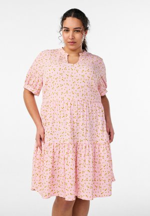 Vrouw draagt een roze jurk tot op de knie met een geel bloemenpatroon, korte pofmouwen en een V-hals, staand tegen een witte achtergrond.