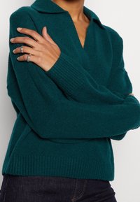 Osoba ve tvídovém svetr s límečkem v teplých tónech, se založenýma rukama, na levé ruce má dvě stříbrné prsteny, v kombinaci s tmavými džíny.