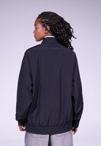 Casaco leve preto com gola alta, ajuste oversized, punhos e barra elásticos. Textura suave, sem padrões visíveis ou detalhes em hardware.