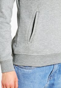 Sweat-shirt gris avec une poche latérale, des poignets et un ourlet côtelés. Composé d'un tissu doux et texturé, il offre une coupe décontractée. Porté avec un jean bleu.