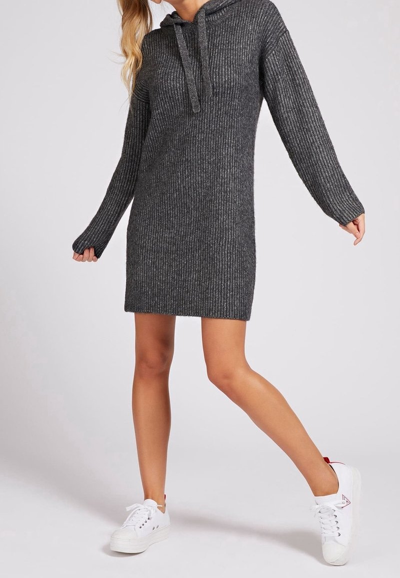 Femme portant une robe à capuche en maille côtelée gris foncé et des baskets blanches, posant avec une jambe tendue et les bras légèrement écartés.