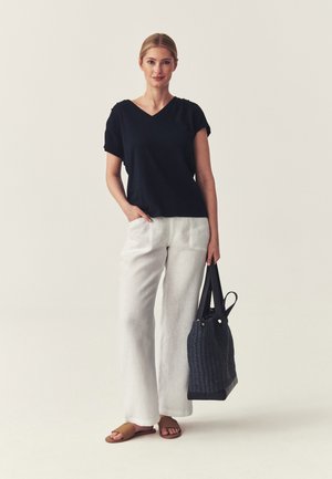 Woman wearing a black V-neck top, white wide-leg pants, tan sandals, holding a black woven tote bag, standing on a plain light background.