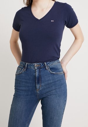 Kobieta ubrana w dopasowany granatowy t-shirt z dekoltem w serek i niebieskie dżinsy z wysokim stanem, z jedną ręką opartą na biodrze.