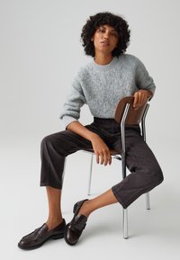 Gray fuzzy sweater with ribbed neckline, dark cropped pants, and brown loafers. Model seated on a chrome chair with wooden seat.