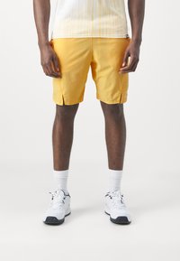 Gelbe Sportshorts mit seitlichen Schlitzen, aus leichtem Material. Kombiniert mit weißen Sneakers und knöchelhohen Socken.