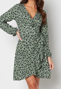 Green floral wrap dress with black patterns, long puff sleeves, V-neckline, and a ruffled hem. Made from textured fabric with a tailored fit.
