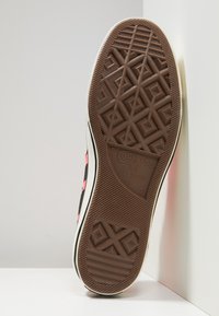 Gummisulor till sneakers med ett brunt, texturerat mönster som innehåller en blandning av geometriska former och en logotyp. Sidan har svarta och rosa accenter.