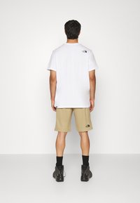Man som bär vit kortärmad t-shirt, beige knälånga shorts, svarta strumpor och svarta sneakers, står vänd mot en enkel vit vägg.