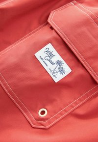 Korallszínű anyag sima textúrával, fehér címkével, amelyen a "West Coast Surf Supply" felirat olvasható, pálmafás grafikákkal és eltérő varrással részletezve.