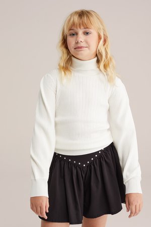 Pulover alb cu guler înalt, cu mâneci lungi, asortat cu o fustă neagră cu talie ornamentată cu paiete. Design simplu, ajustat și texturat.