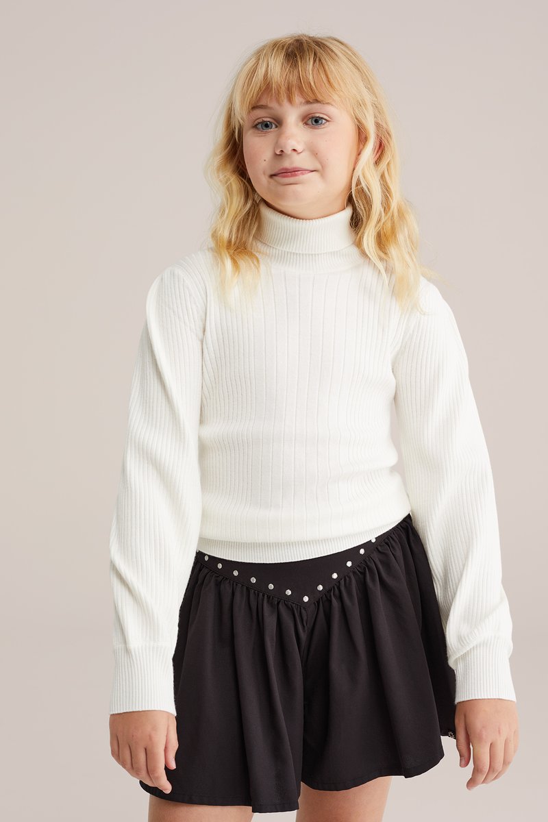 Bela rebrasta turtleneck pulover z dolgimi rokavi, v kombinaciji s črno krilo s pasom iz bleščic. Enostaven dizajn, prilegnjen in teksturiran.