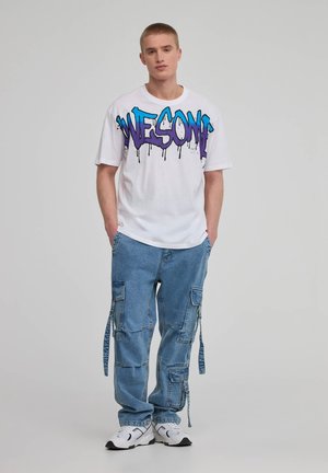 T-shirt bianco a maniche corte con una grafica multicolore "AWESOME". Indossato con pantaloni cargo blu chiaro dotati di tasche e dettagli legati.