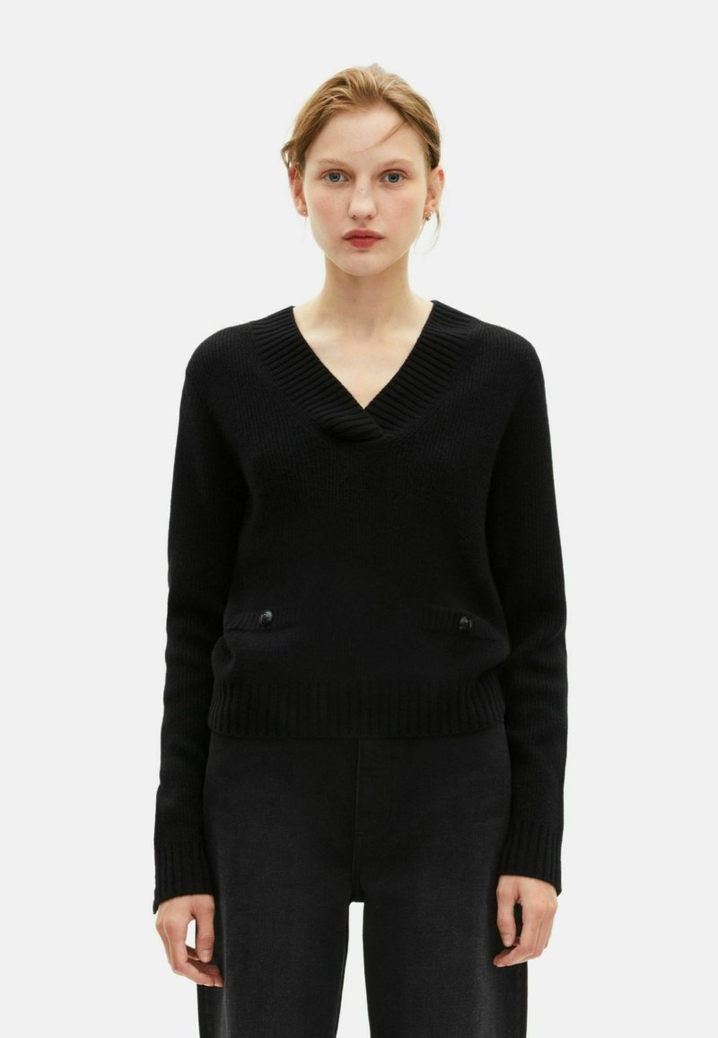 Zwarte gebreide V-hals trui met ribbelstructuur, voorzien van twee zakken aan de voorkant en knopenaccenten. Gecombineerd met donkere high-waisted broeken.