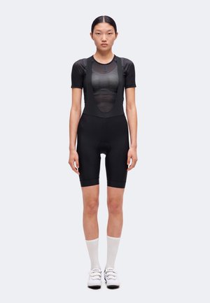Femme debout portant un cuissard de cyclisme noir avec bretelles, un haut noir en maille à manches courtes, des chaussettes blanches et des chaussures de cyclisme blanches.