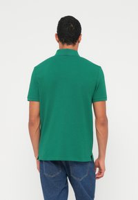 Πράσινο πόλο μπλουζάκι με γιακά και κοντά μανίκια, κατασκευασμένο από λείο ύφασμα· η πίσω όψη δείχνει ίσια κοπή με πλευρικές σχισμές.