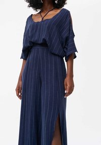 Femme portant un pantalon large à fines rayures bleu marine avec des fentes et un haut ample assorti avec un détail d'épaule découpée.