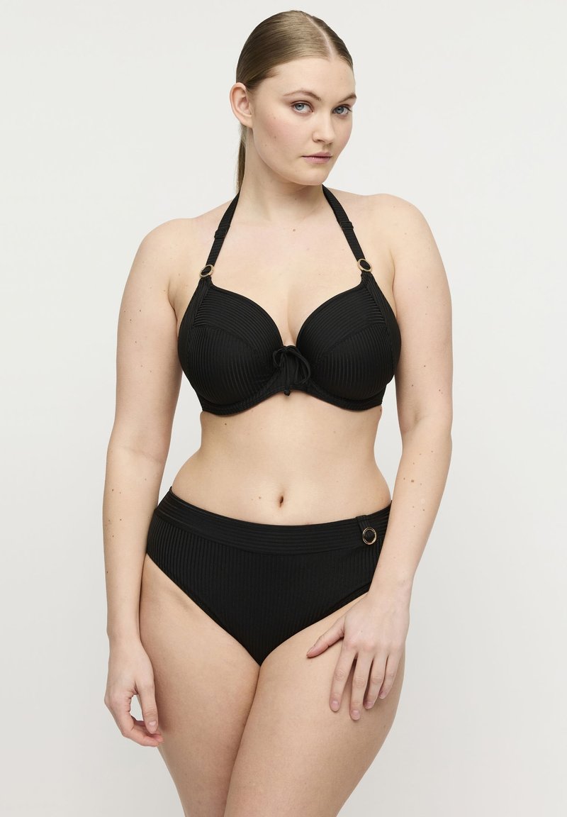 Kobieta o jasnej karnacji i związanych do tyłu włosach, ubrana w czarny prążkowany bikini na szyję z złotymi detalami w postaci pierścieni, stojąca na tle jednolitego jasnego tła.