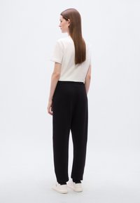 Λευκό βαμβακερό t-shirt με κοντά μανίκια, συνδυασμένο με μαύρο παντελόνι φόρμας με ελαστική μέση και λευκά αθλητικά παπούτσια, σε προβολή από πίσω.