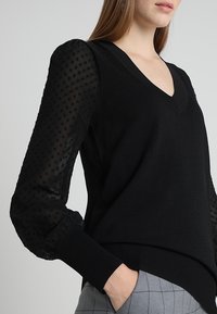 Musta V-kaulusvetoketju, jossa on läpikuultavat pallohihat mustilla pilkuilla. Resorireunukset hihoissa ja helmassa. Valmistettu pehmeästä, teksturoidusta materiaalista.