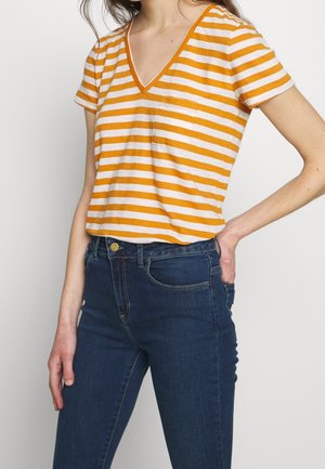 Kobieta ubrana w żółto-biały T-shirt w paski z dekoltem w serek i krótkimi rękawami oraz ciemnoniebieskie, wysokie spodnie jeansowe na gładkim tle.