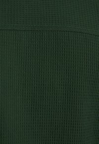 Zelená texturovaná látka se vzorem mřížky, zobrazující vzor vaflového pletení. Povrch působí měkce a mírně vystouplý.