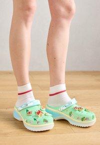 Beine in weißen Socken mit roten Streifen und grünen Plateauschuhen, die mit bunten Cartoon-Charms verziert sind, auf einem Holzboden.