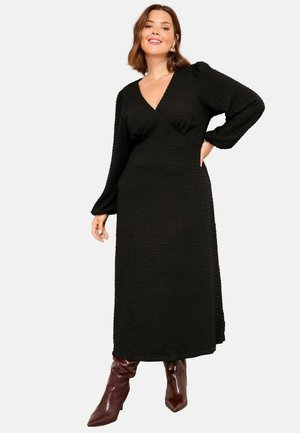 Naeratav naine, kes kannab pikka musta tekstuuriga V-kaelusega kleiti, millel on volangvarrukad, ning pruunide terava ninaga kontsakingi, poseerib ühe käega puusal.