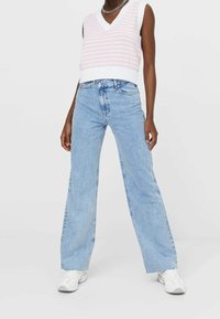 Witte mouwloze trui met een roze geometrisch patroon, gecombineerd met lichtblauwe high-waisted wijde jeans en witte sneakers. Accenten van zilveren ketting.