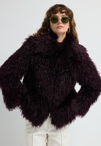 Kvinde iført en mørk lilla fluffy faux fur-jakke, hvide bukser, guld hoops-øreringe og grøntonede solbriller mod en ensfarvet baggrund.