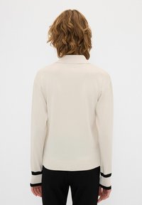Maglione crema con maniche lunghe, polsini a coste con strisce nere e colletto classico. Texture liscia e design aderente.