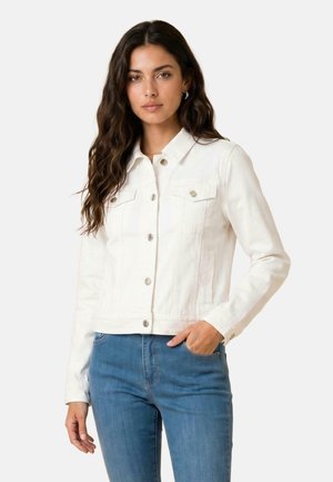 Vrouw met lang donker haar draagt een witte jas met knopen en blauwe jeans, staand met één hand in haar zak tegen een effen achtergrond.