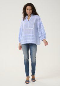 Blå och vit randig blus med hög krage, överdimensionerade ärmar och plisserad design, i kombination med figursydda blå jeans och öppna klackskor.